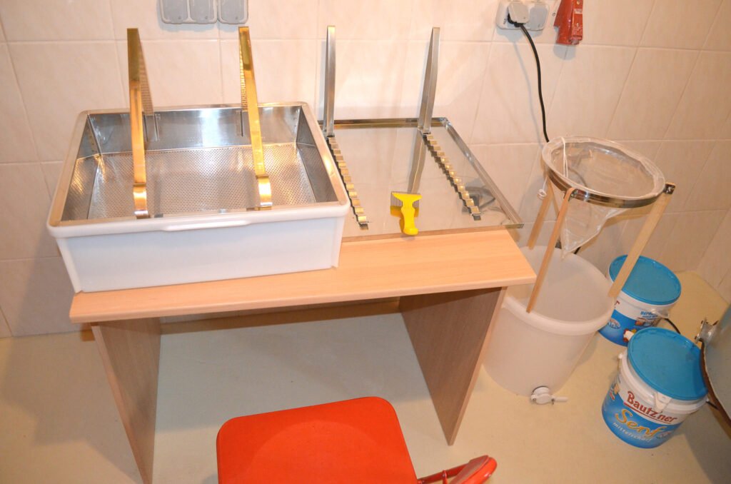 A conical honey strainer sitting in a stand above a food-grade bucket with a gate, beside an uncapping tray