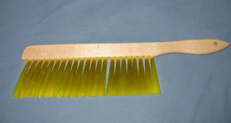 Bee brush with a light wooden handle and long, soft yellow bristles photographed on a plain blue background