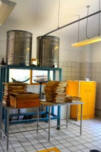 Two tall stainless honey settling tanks raised on a steel frame with a tap, above stacked empty frames