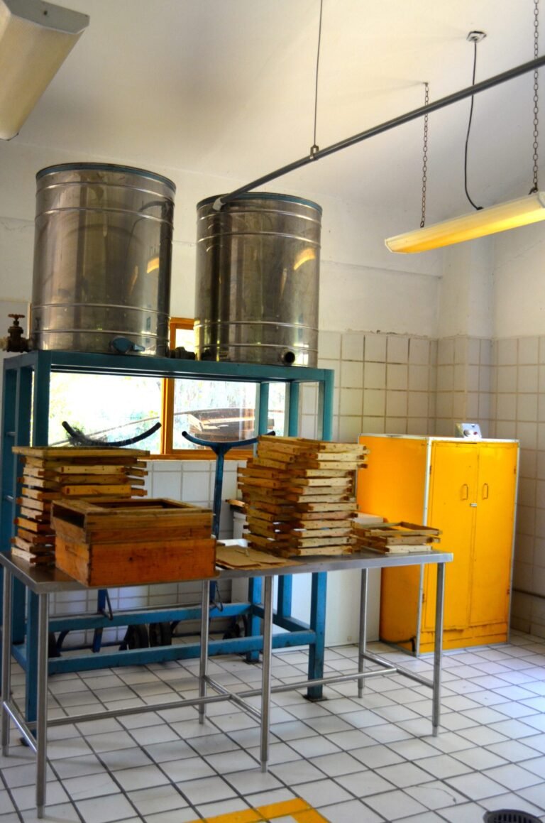 Two tall stainless honey settling tanks raised on a steel frame with a tap, above stacked empty frames