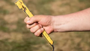 Close-up of a beekeeper's hand gripping a worn yellow hive tool with a hooked end, used to pry boxes and frames apart