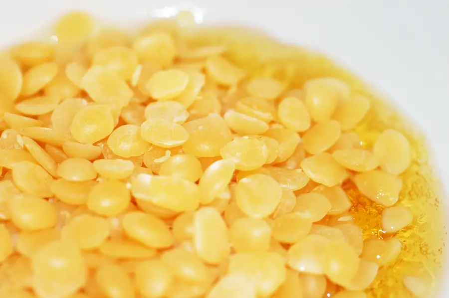 Yellow beeswax pellets melting into a shallow pool of clear liquid wax in a white bowl