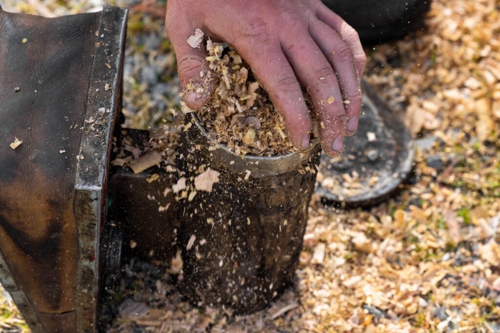 Hand dropping a loose handful of dry wood shavings into the open firebox of a bee smoker