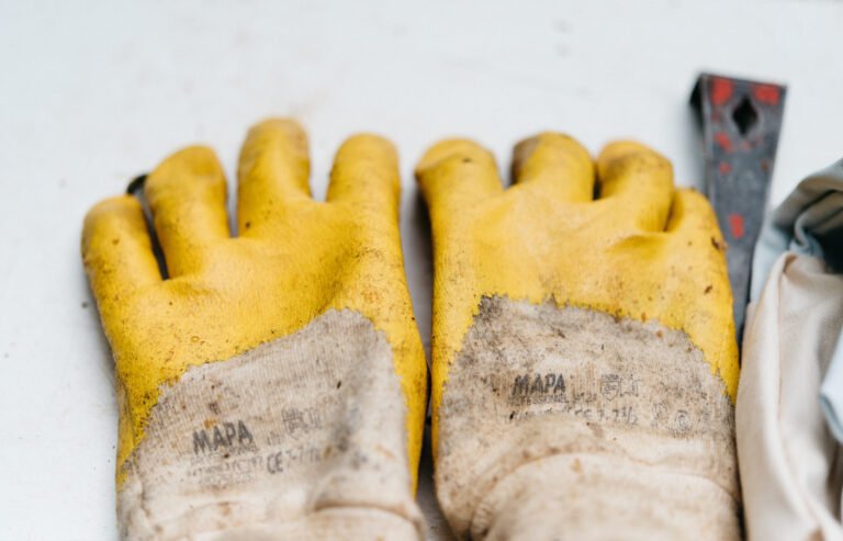 Worn yellow rubber-coated work gloves beside a hive tool and a bee suit