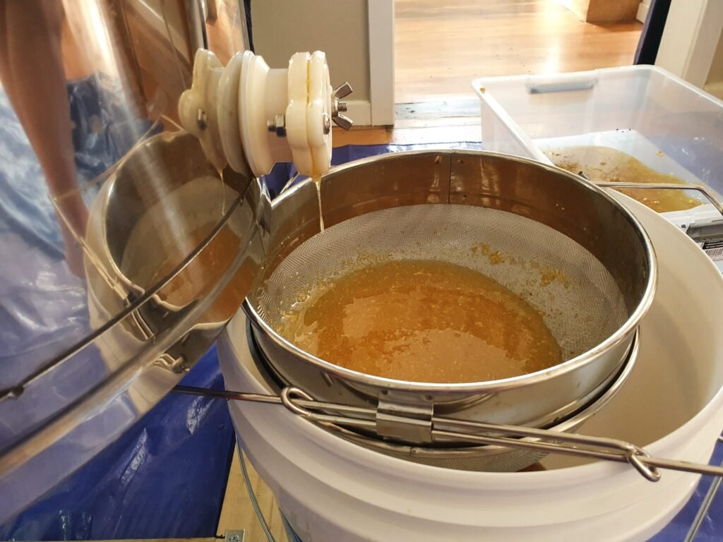 Photo of honey running from an extractor gate into a double stainless steel strainer over a bucket, with foam on top