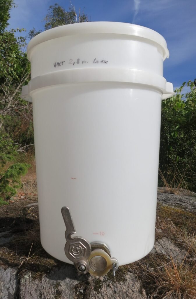 Photo of a tall white plastic honey settling tank with a metal honey gate, holding about 13 gallons