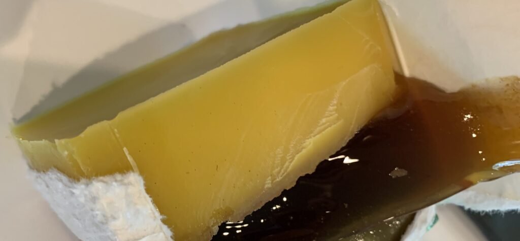 Photo of a set cake of rendered beeswax in a cut-open carton, with dark liquid and residue settled beneath it
