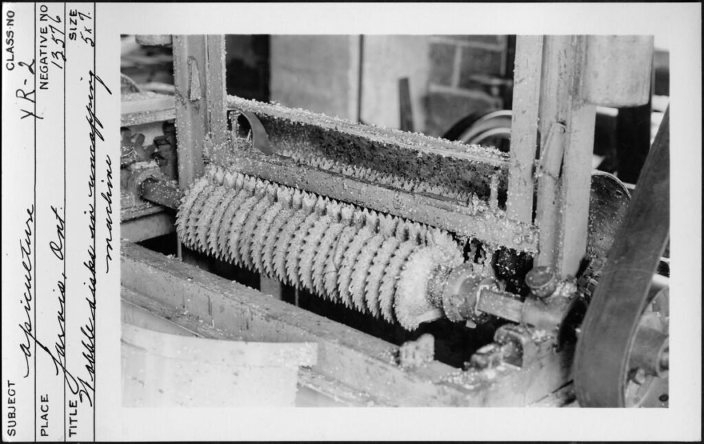 Archival photograph of a mid-20th-century wobble-disk uncapping machine at an Ontario apiary, an early mechanized ancestor of today's automated Cowen uncapper