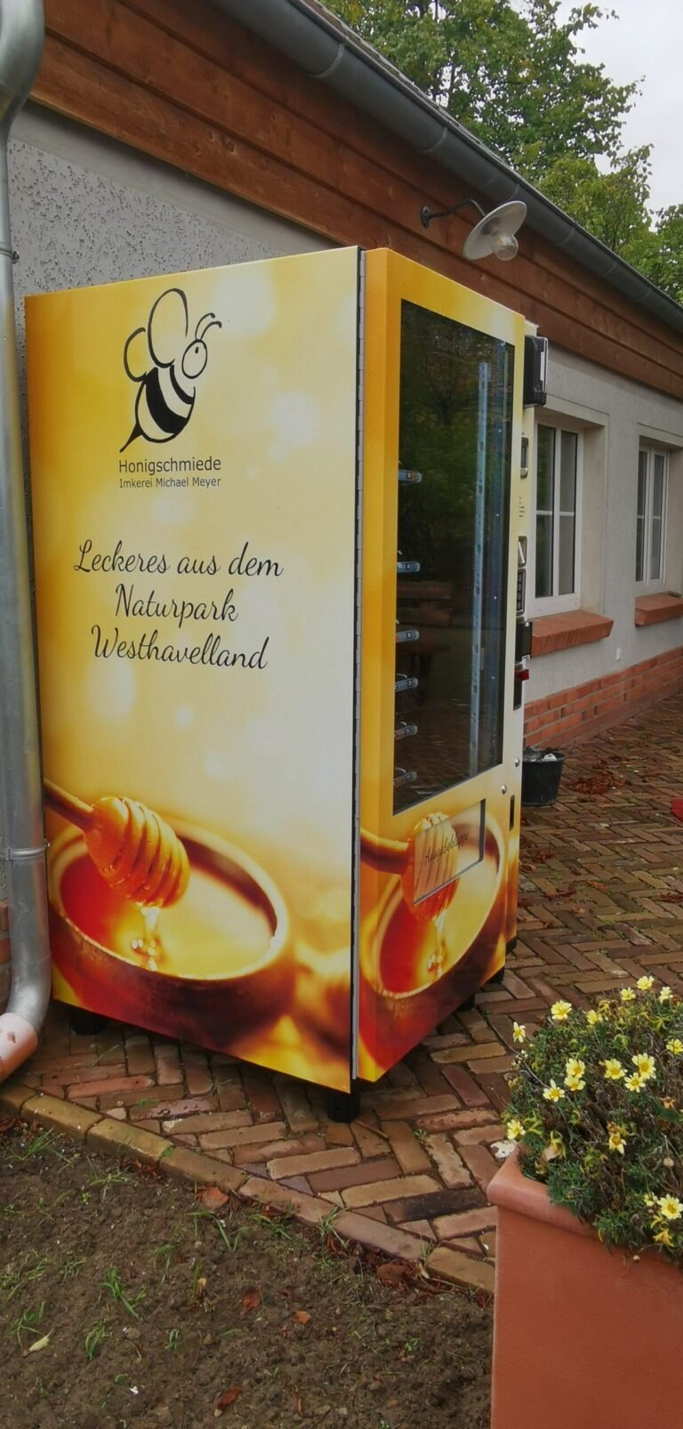 A coin-operated honey vending machine (Honigautomat) mounted at a German farm gate, selling jars of local honey around the clock