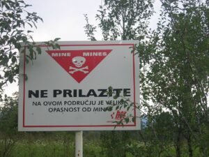 A red and white minefield warning sign reading NE PRILAZITE (Do Not Approach) with a skull and crossbones, posted in rural Croatia