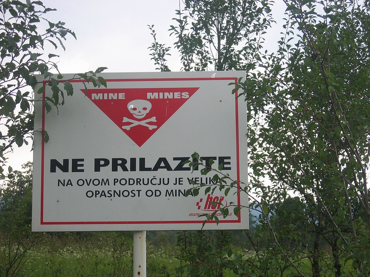 A red and white minefield warning sign reading NE PRILAZITE (Do Not Approach) with a skull and crossbones, posted in rural Croatia