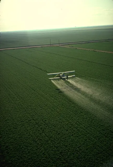 Crop duster airplane spraying pesticide over a large agricultural field