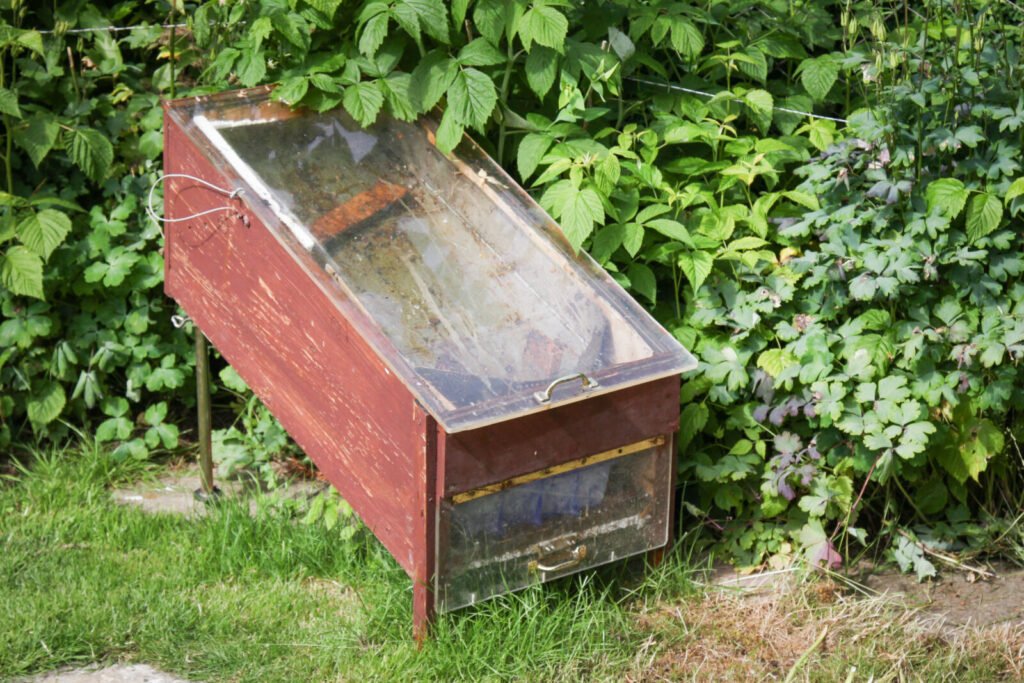 A home-built red wooden solar wax melter with a glass lid and a glass-fronted collection drawer beneath the tray