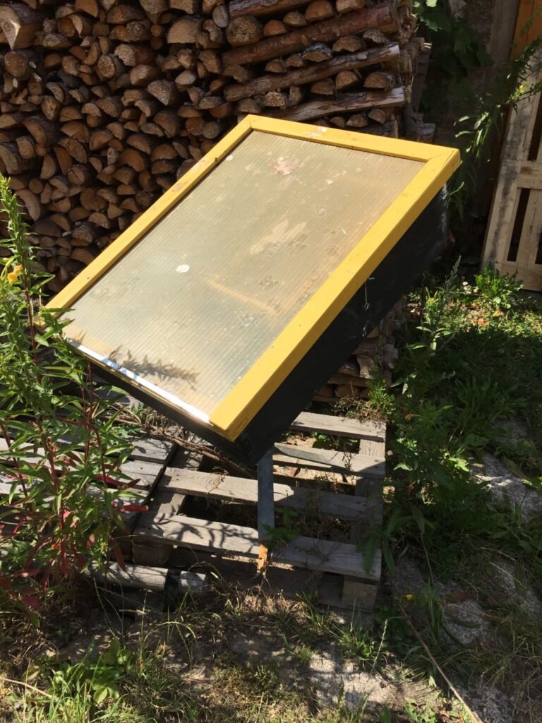 A black-painted wooden solar wax melter with a yellow-framed glazed lid, tilted toward the sun on a pallet stand