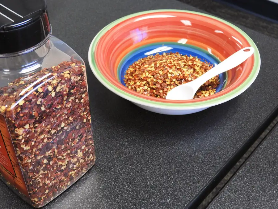 Dried red pepper flakes in a clear plastic spice jar and a colorful bowl with a small white spoon on a dark counter