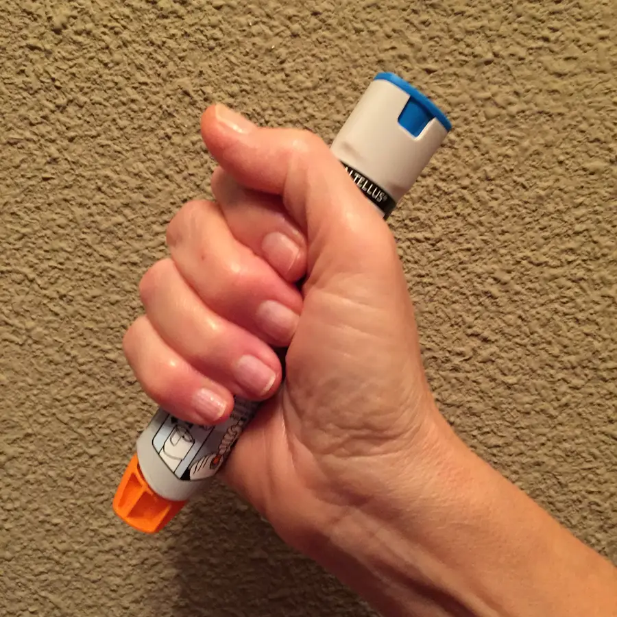 Hand gripping an epinephrine auto-injector in a fist with the orange needle end pointing down and blue safety cap on top
