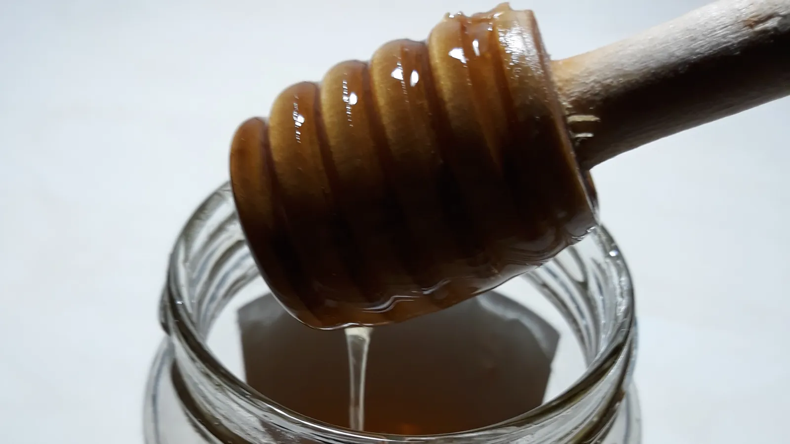 Wooden honey dipper dripping a thin stream of thick amber honey into an open glass jar