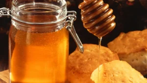 Glass jar of liquid honey beside a wooden dipper dripping honey onto a biscuit